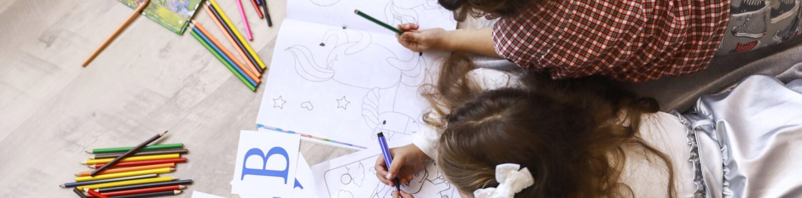 Top view of two tiny girls which are drawing in the coloring boo Top view of two tiny girls which are drawing in the coloring book laying on the floor on the blanket